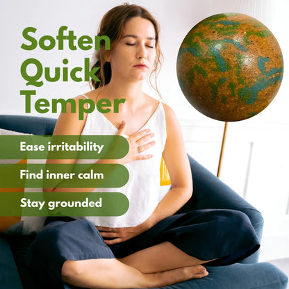 A woman sits cross-legged on a blue couch, holding her chest as if in relief, showing bracelet softens quick temper ease irritability, find inner calm, and stay grounded.